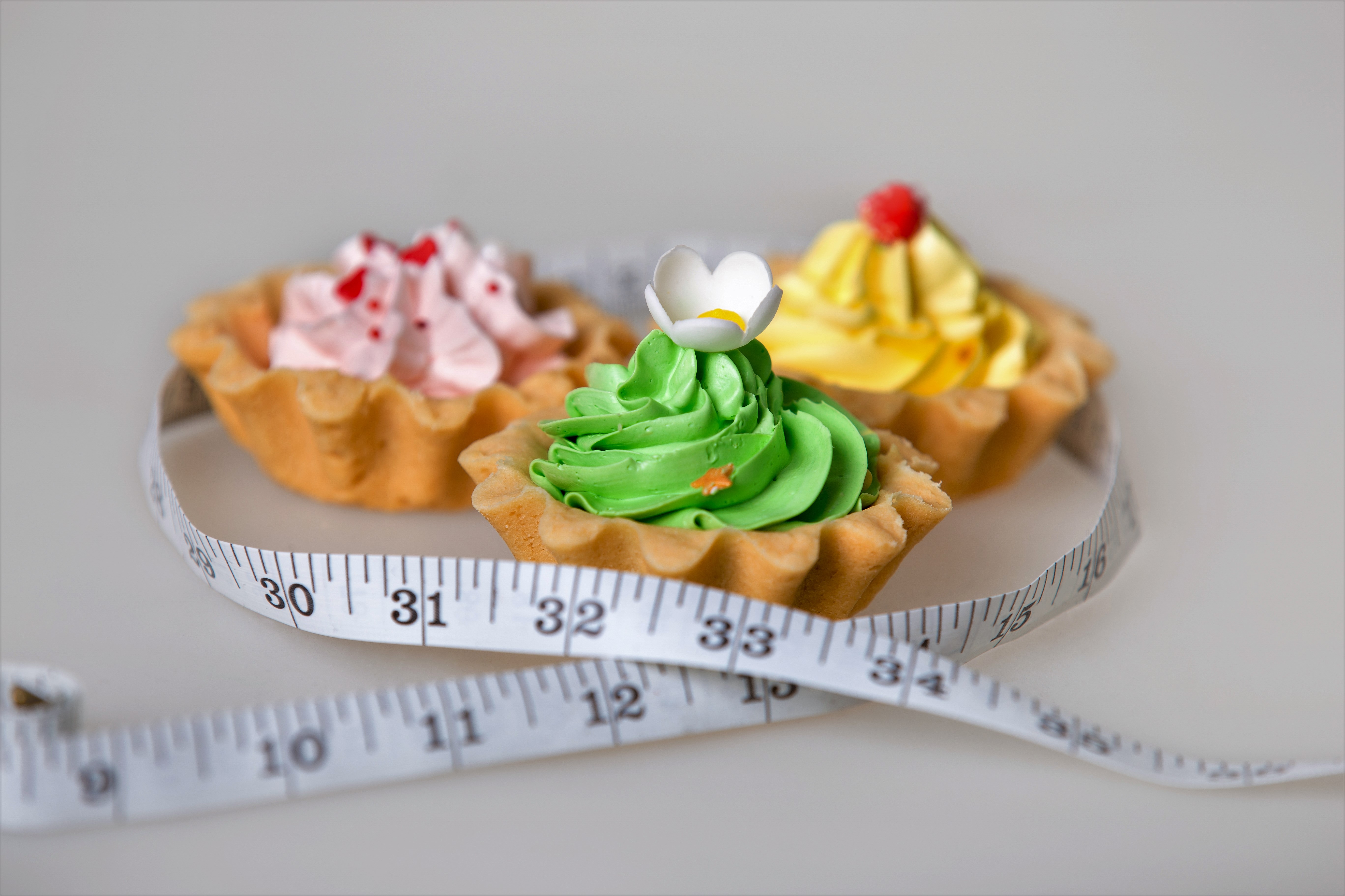 Three colorful green, pink and yellow tart cakes wrapped in measuring tape on white background, unhealthy lifestyle concept, studio